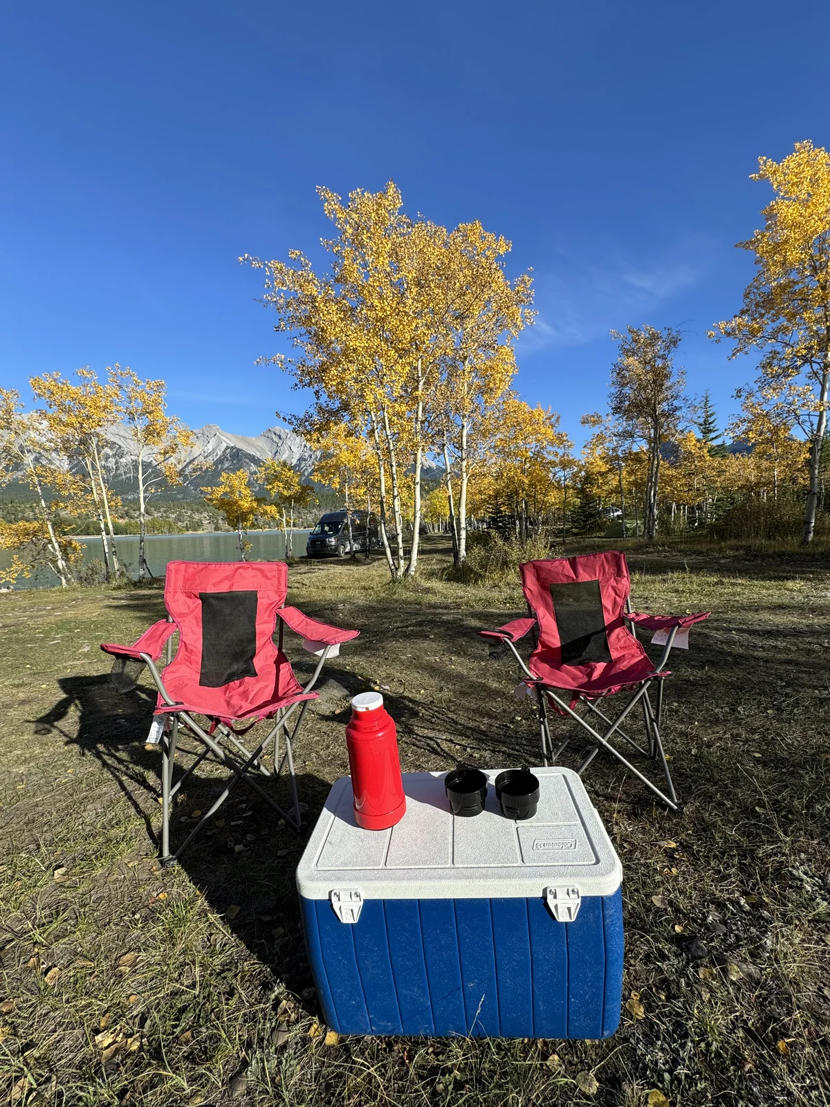 Camp chairs, a cooler, and a hot thermos set up at a fall campsite beside a mountain lake
